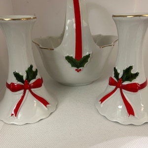 Puede incluir: Un juego de tres candelabros de Navidad de porcelana blanca con decoraciones de acebo rojo y verde y un lazo. El candelabro del centro es una cesta con un asa de cinta roja.
