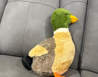 Könnte beinhalten: Ein Plüsch-Entenspielzeug mit grünem Kopf, braunem und braunem Körper und orangefarbenen Füßen. Die Ente liegt auf einem grauen Sofa.