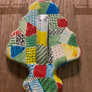 May include: A vintage metal cookie cutter shaped like a Christmas tree. The cutter is painted white with a patchwork design of red, green, blue, yellow, and black.