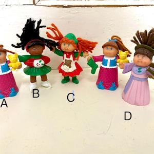 May include: Five vintage Cabbage Patch Kids figurines. Each doll has a unique outfit and hairstyle. The dolls are holding accessories like a teddy bear, a green sock, and a horn. The dolls are in various colors, including pink, green, and red.