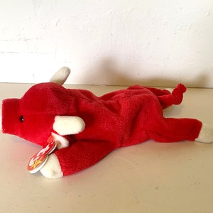 May include: Red plush bull Beanie Baby with white horns and a white tag that says "Beanie Baby".
