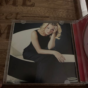 May include: A woman with blonde hair is sitting on a white chair. She is wearing a black dress and has her head resting on her hand. The background is a white wall.