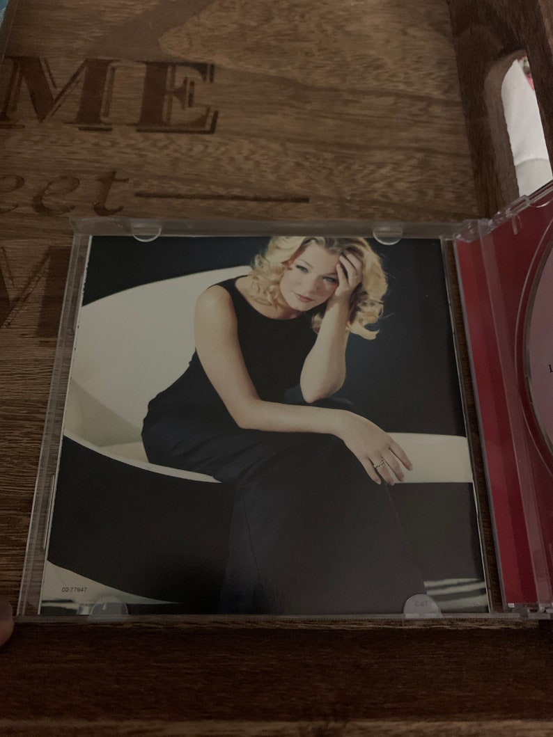 May include: A woman with blonde hair is sitting on a white chair. She is wearing a black dress and has her head resting on her hand. The background is a white wall.