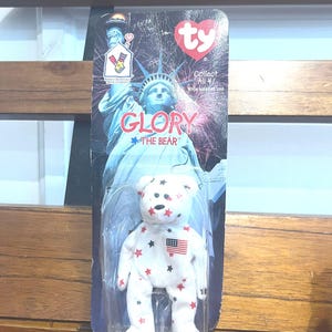 Puede incluir: Un oso de peluche blanco Ty Beanie Baby llamado "Glory the Bear" con estrellas rojas y una pequeña bandera estadounidense en el pecho. El oso está empaquetado en una bolsa de plástico con una etiqueta Ty que muestra la Estatua de la Libertad y las palabras "Collect All 4!"