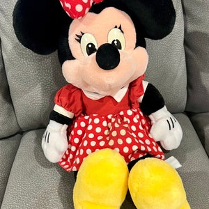 K&ouml;nnte beinhalten: Ein Pl&uuml;schtier von Minnie Mouse, das ein rotes Kleid mit wei&szlig;en Punkten und eine rote Schleife mit wei&szlig;en Punkten tr&auml;gt. Das Spielzeug hat schwarze Ohren, ein rosa Gesicht und gelbe Schuhe.