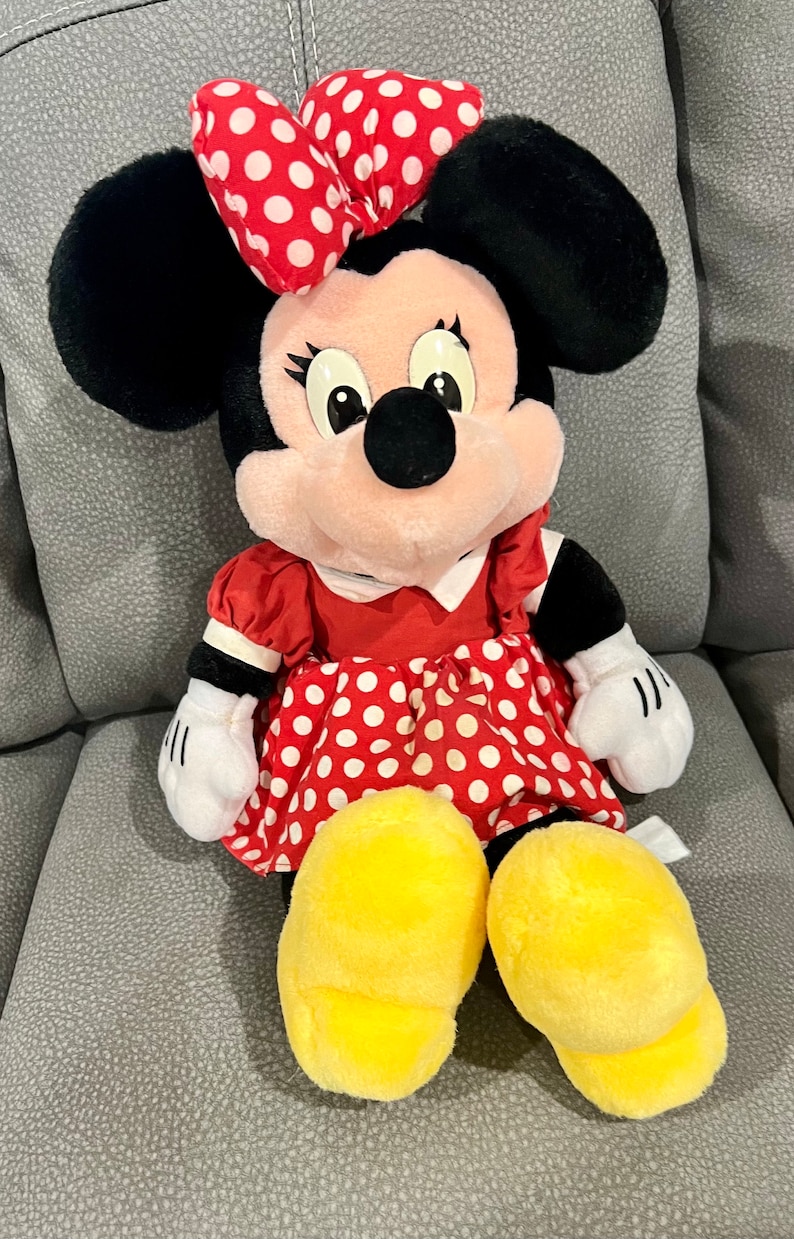 K&ouml;nnte beinhalten: Ein Pl&uuml;schtier von Minnie Mouse, das ein rotes Kleid mit wei&szlig;en Punkten und eine rote Schleife mit wei&szlig;en Punkten tr&auml;gt. Das Spielzeug hat schwarze Ohren, ein rosa Gesicht und gelbe Schuhe.