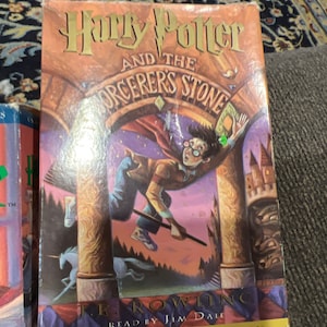 May include: A hardcover copy of "Harry Potter and the Sorcerer's Stone" by J.K. Rowling. The cover features an illustration of Harry Potter flying on a broomstick. The title is in gold lettering, with the author's name and "An Unabridged Production" at the bottom.