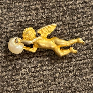 Könnte beinhalten: Eine goldene Cherubfigur mit Flügeln, die eine weiße Perle hält. Der Cherub fliegt mit ausgebreiteten Flügeln.