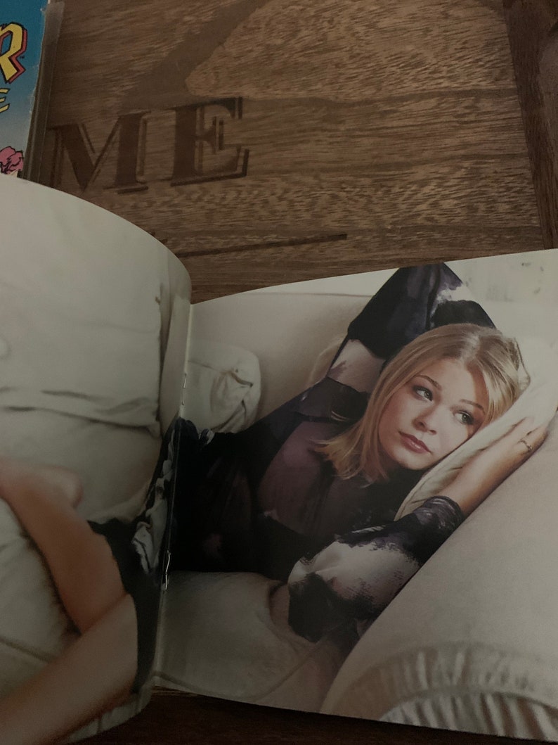 May include: A woman with blonde hair is lying on a white bed with a white pillow. She is wearing a black and white patterned shirt.