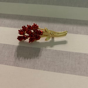 May include: A gold-toned brooch with a bouquet of red enamel flowers. The flowers are arranged in a cluster and have a slightly textured finish.