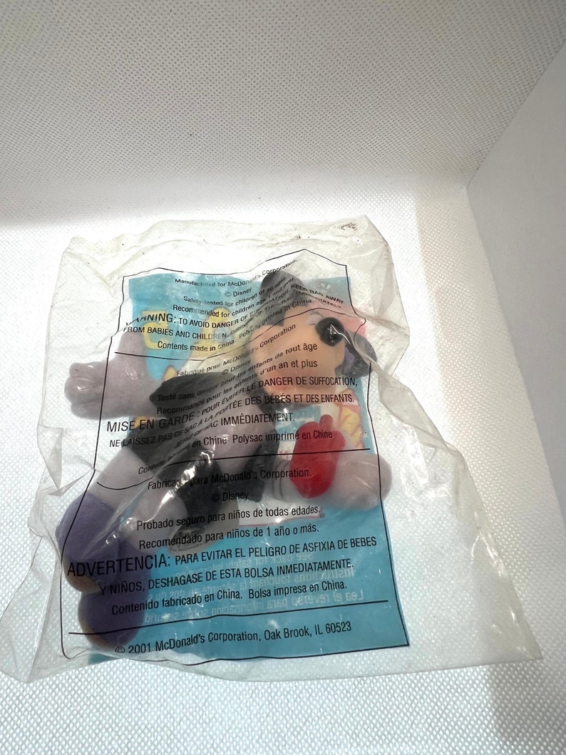 Puede incluir: Un personaje de juguete de pl&aacute;stico morado, rojo y amarillo con una etiqueta de advertencia en espa&ntilde;ol e ingl&eacute;s. La etiqueta dice "ADVERTENCIA: PARA EVITAR EL PELIGRO DE ASFIXIA DE BEBES Y NI&Ntilde;OS, DESHAGASE DE ESTA BOLSA INMEDIATAMENTE. Contenido fabricado en China. Bolsa impresa en China." La etiqueta tambi&eacute;n incluye el texto "Probado seguro para ni&ntilde;os de todas edades. Recomendado para ni&ntilde;os de 1 a&ntilde;o o m&aacute;s."