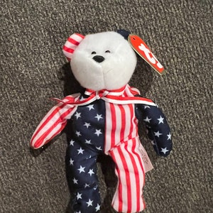 May include: A white plush bear wearing a red, white, and blue striped and star-patterned outfit. The bear has a black nose and black eyes. The bear is holding a red tag with the word "TY" on it.