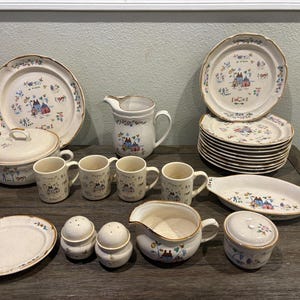 Może przedstawiać: Kolekcja kremowych ceramicznych naczyń z złotym obramowaniem i fantazyjnym wzorem wioski. Zawiera talerze, miski, kubki, dzbanek do śmietanki, półmisek do serwowania oraz solniczki i pieprzniczki. Zestaw jest ułożony na drewnianej powierzchni.