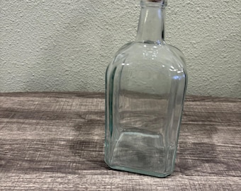 Könnte beinhalten: Eine klare Glasflasche mit einem Korkverschluss. Die Flasche ist leer und hat eine quadratische Form.