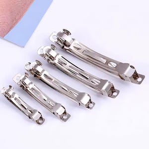 May include: A collection of silver-colored metal hair barrettes in various sizes. The barrettes are arranged diagonally, showcasing their sleek, rectangular design with a hinged clasp and decorative details. The background is a soft, neutral color.