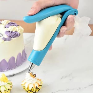 Könnte beinhalten: Eine blaue Spritztülle wird verwendet, um einen Cupcake mit weißem Zuckerguss zu dekorieren. Die Spritztülle wird von einer Hand gehalten und ist mit weißer Creme gefüllt. Ein glasierter Kuchen und andere Cupcakes sind im Hintergrund zu sehen.