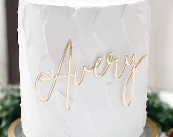 Può includere: Una torta bianca glassata con il nome "Avery" in oro. La torta ha una superficie strutturata e poggia su un alzatina dorata. La torta è un dolce per una festa.