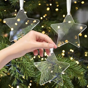 Könnte beinhalten: Transparente sternförmige Weihnachtsschmuckstücke mit weißen Bändern. Die Ornamente hängen an einem grünen Weihnachtsbaum mit gelben Lichtern im Hintergrund. Eine Hand hält einen der Sterne.
