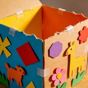 May include: A handmade cardboard box with colorful decorations. The box features a blue side with a yellow star, red rectangle, and orange dog shape. The other sides are decorated with a purple hexagon, yellow giraffe, and pink flower.