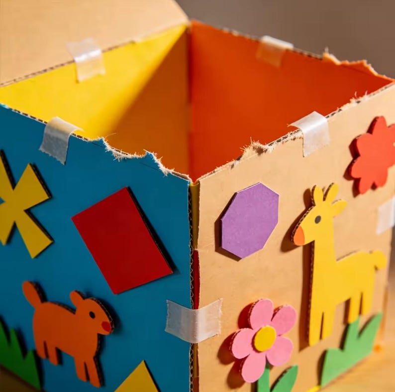 May include: A handmade cardboard box with colorful decorations. The box features a blue side with a yellow star, red rectangle, and orange dog shape. The other sides are decorated with a purple hexagon, yellow giraffe, and pink flower.