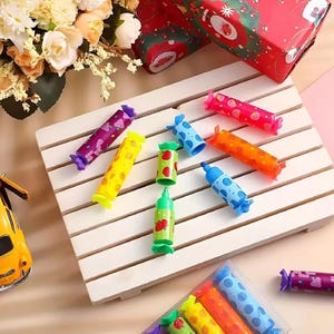 以下が含まれることがあります： 紫、黄色、オレンジ、青、ピンクなど、さまざまな色のカラフルなキャンディー型のペン。果物のデザインや水玉模様などがあります。白い木の表面に配置されています。