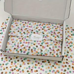 Puede incluir: Una caja de envío de cartón blanco que contiene artículos envueltos en papel de seda translúcido con un patrón de lunares multicolor. Una pegatina blanca rectangular con un borde decorativo y el texto Thank you está centrada en el papel.