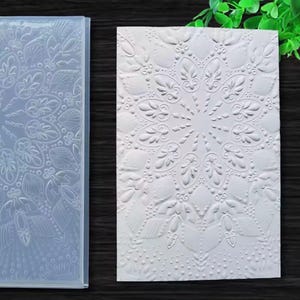 Könnte beinhalten: Ein rechteckiger Prägefolder aus transparentem Kunststoff mit einem detaillierten Mandala-Muster aus symmetrischen Blumen- und Geometrieformen, neben einem weißen Kartonblatt mit demselben eingeprägten radialen Design.