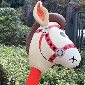 May include: Inflatable toy horse head with a cream-colored face, brown mane, and red and pink bridle. The horse has a black cross for a mouth and is designed for children's play.