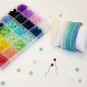 Könnte beinhalten: Ein klarer Kunststoff-Organizer gefüllt mit bunten runden Perlen in verschiedenen Farbtönen, darunter Lila, Rosa, Blau, Grün und Gelb. Mehrere Armbänder mit Perlen in Blau- und Grüntönen werden auf einem weißen Kissen präsentiert. Ein Paar rote und silberne Ohrringe sind ebenfalls zu sehen.