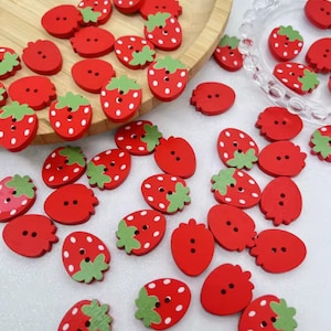 Könnte beinhalten: Eine Sammlung roter Holzknöpfe in verschiedenen Formen, darunter Erdbeeren und runde Knöpfe. Die Erdbeerknöpfe haben grüne Blätter und weiße Punkte. Einige Knöpfe liegen auf einem Holztablett und einer durchsichtigen Schale.