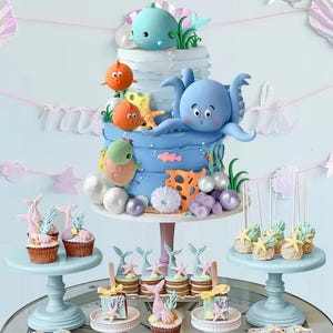 以下が含まれることがあります： 海のテーマのマルチティアケーキとカップケーキ。ケーキは青色で、クジラ、タコ、魚、カメなどの海の生き物の装飾が施されています。カップケーキとケーキポップは、人魚の尾とヒトデで飾られています。