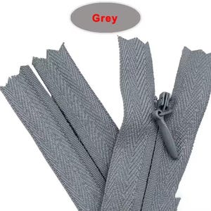 Puede incluir: Primer plano de varias cremalleras grises, con un tirador visible. La palabra "Grey" se muestra en un &oacute;valo gris encima de las cremalleras. Las cremalleras tienen un patr&oacute;n de espiga.