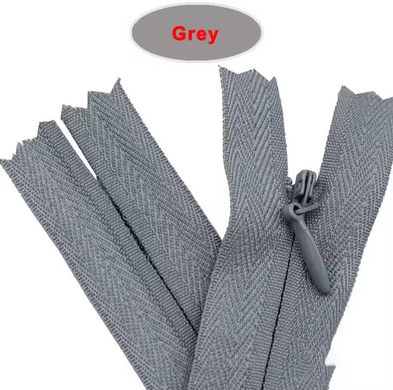 Puede incluir: Primer plano de varias cremalleras grises, con un tirador visible. La palabra "Grey" se muestra en un &oacute;valo gris encima de las cremalleras. Las cremalleras tienen un patr&oacute;n de espiga.