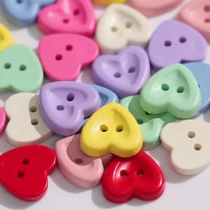 May include: A selection of heart-shaped buttons in pastel shades such as pink, yellow, green, and purple. Each button features two holes for sewing. The buttons are scattered, forming a colourful and playful arrangement.