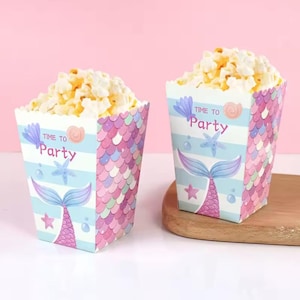 May include: Two mermaid-themed popcorn boxes filled with popcorn. The boxes feature a mermaid tail design, seashells, and the text "Time to Party." The boxes have a blue, pink, and purple colour scheme.