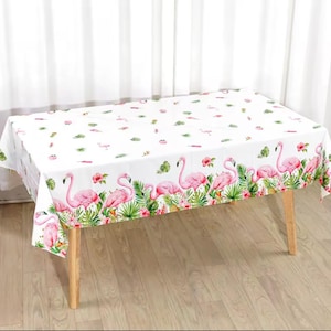 May include: White rectangular tablecloth featuring a repeating pattern of pink flamingos, green tropical leaves, and pink flowers. The border showcases a row of flamingos amidst lush foliage. The tablecloth is set on a wooden table.