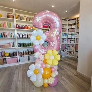 Peut inclure: Un grand ballon rose en forme de chiffre 3, orné de marguerites blanches et d'une écriture dorée. Il est entouré d'un ensemble de ballons blancs, jaunes et roses de différentes tailles, créant une présentation festive. Parfait pour un anniversaire.
