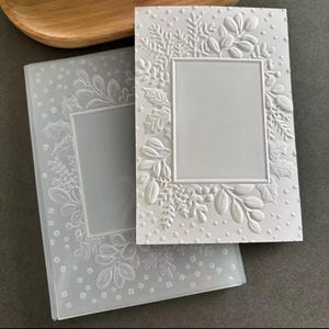 Puede incluir: Dos marcos de fotos rectangulares blancos. Uno está grabado con un diseño floral y de copo de nieve. El otro es un marco transparente con un diseño en relieve similar. Ambos tienen una abertura rectangular para una foto.