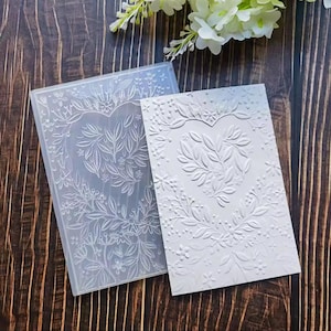Könnte beinhalten: Ein klarer, rechteckiger Prägeordner mit einem herzförmigen Blumendesign, neben einer weißen geprägten Karte mit demselben Muster. Das Design zeigt Blätter und kleine Blüten. Auf einer dunklen Holzoberfläche.