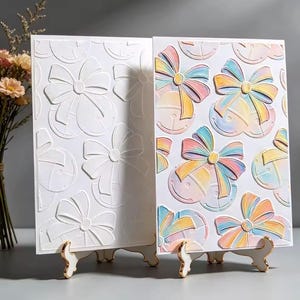Op de afbeelding: Twee rechthoekige kaarten op witte standaards. De linkerkaart heeft een wit reliëfpatroon van strikken en bellen. De rechterkaart toont hetzelfde reliëfpatroon met een veelkleurige pastelkleurige gradiëntafwerking in roze, blauw en geel.