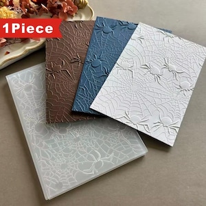 May include: Four rectangular embossing folders featuring a raised spider web and spider pattern. The set includes one translucent, one brown, one dark blue, and one white folder. Each folder displays a repeating design of arachnids and webs.