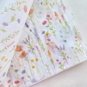 Peut inclure: Tissu blanc avec un motif floral à l'aquarelle. Le motif présente des fleurs sauvages colorées dans des tons de rose, de violet, d'orange et de jaune, avec des tiges et des feuilles vertes. Le tissu est plié, révélant le motif.