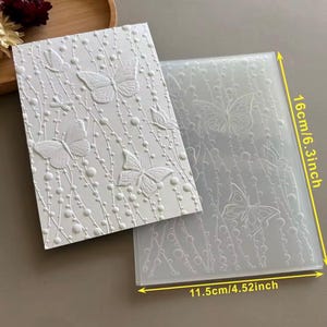 May include: A white rectangular embossing folder featuring a raised butterfly and beaded vine pattern, paired with a translucent plastic template. The set measures 6.3 inches in height and 4.52 inches in width, showing a detailed butterfly design.