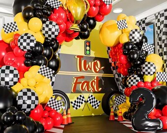 May include: A festive party decoration featuring a yellow backdrop with the words "Two Fast" in red. The scene is adorned with red, black, and yellow balloons, checkered flags, and a gold trophy. A large black number "2" balloon is also present.