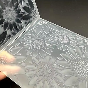 May include: A clear plastic embossing folder featuring a raised sunflower pattern. The rectangular tool has a hinged design with a detailed floral texture on the interior surfaces, designed to create impressions of multiple sunflowers and leaves.