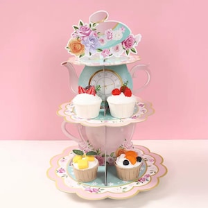 Puede incluir: Un soporte para cupcakes de tres niveles con forma de tetera, adornado con decoraciones florales y cupcakes. El soporte es de color rosa pastel y verde azulado, con un diseño de esfera de reloj. Se exhiben cupcakes con cobertura de frutas.