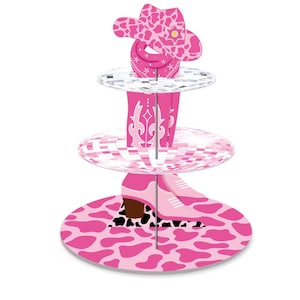 May include: A three-tiered pink and white cowboy-themed cupcake stand. The top tier features a cowboy hat, the middle tier has a disco ball design, and the bottom tier has a cow print design. The stand is designed to hold cupcakes or other small treats.