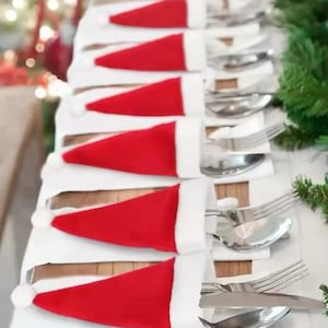 May include: Christmas table setting with silverware in red Santa hat holders with white trim. The cutlery, including forks, spoons, and knives, is arranged on white napkins, ideal for a festive Christmas meal.