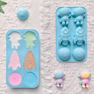 May include: Two light blue silicone moulds featuring space-themed cavities in the shapes of astronauts, rockets, and planets. One mould is filled with colourful liquid, while the other is empty. Two small, solid astronaut figures sit nearby.