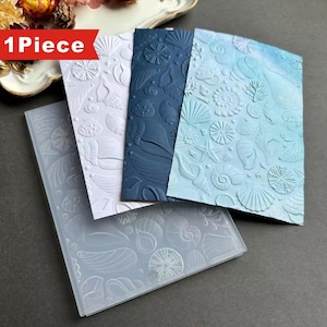 May include: A rectangular plastic embossing folder featuring a raised seashell and starfish pattern, shown with three sample cardstock sheets in white, dark blue, and light blue, each displaying the embossed nautical design and the text 1 Piece.